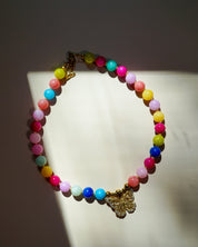 A colorful beaded necklace with pastel and bright round beads, a gold-tone butterfly charm, and a clasp, displayed on a white surface with dramatic shadows.