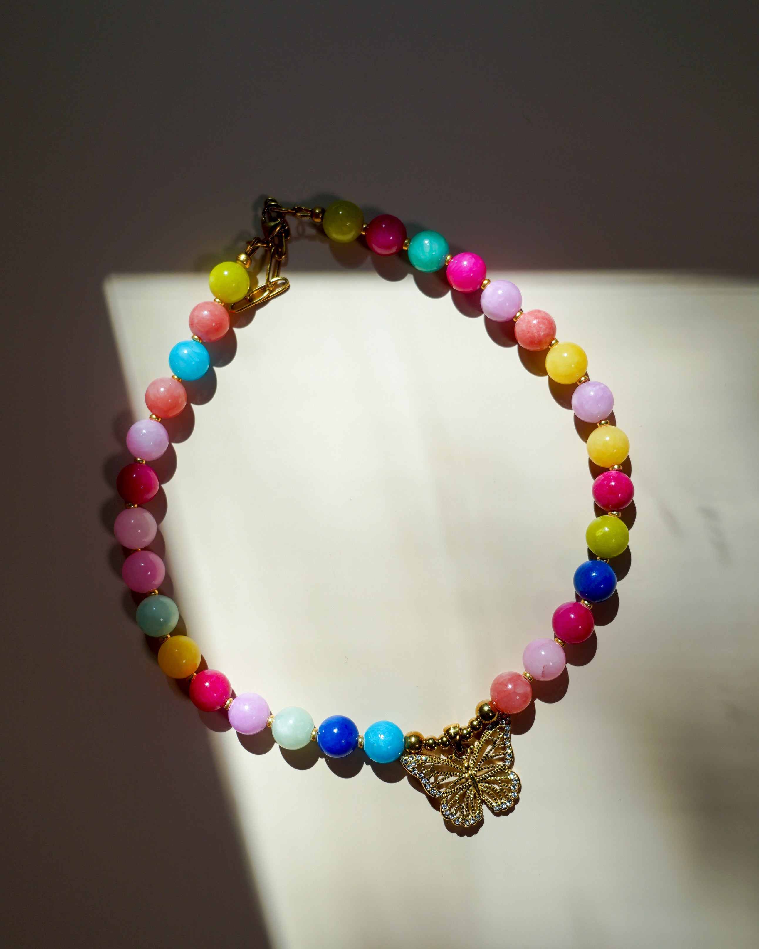 A colorful beaded necklace with pastel and bright round beads, a gold-tone butterfly charm, and a clasp, displayed on a white surface with dramatic shadows.