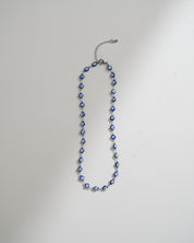 A beaded necklace with blue and white evil eye charms on a silver-toned chain, laid flat on a light gray background.