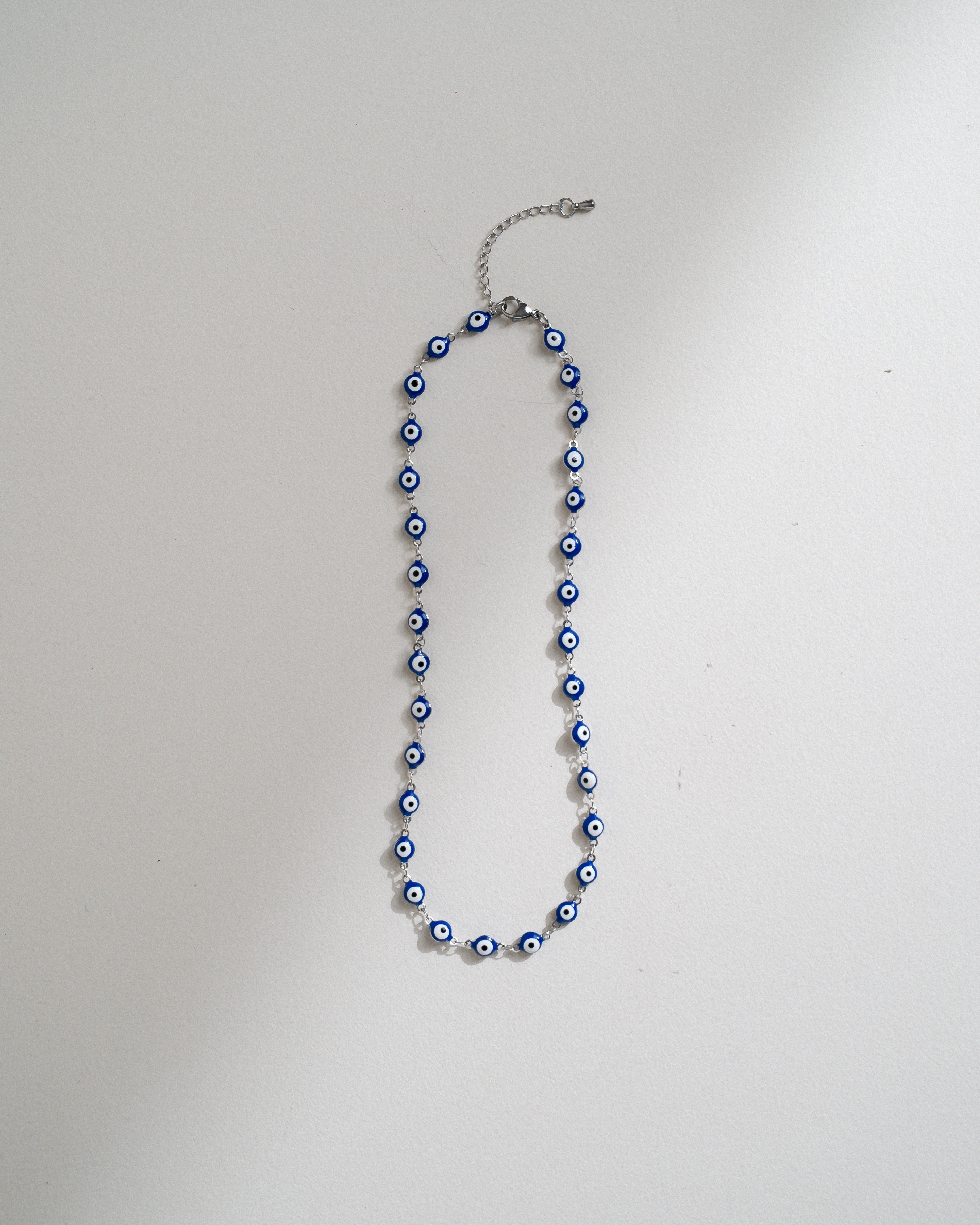 A beaded necklace with blue and white evil eye charms on a silver-toned chain, laid flat on a light gray background.