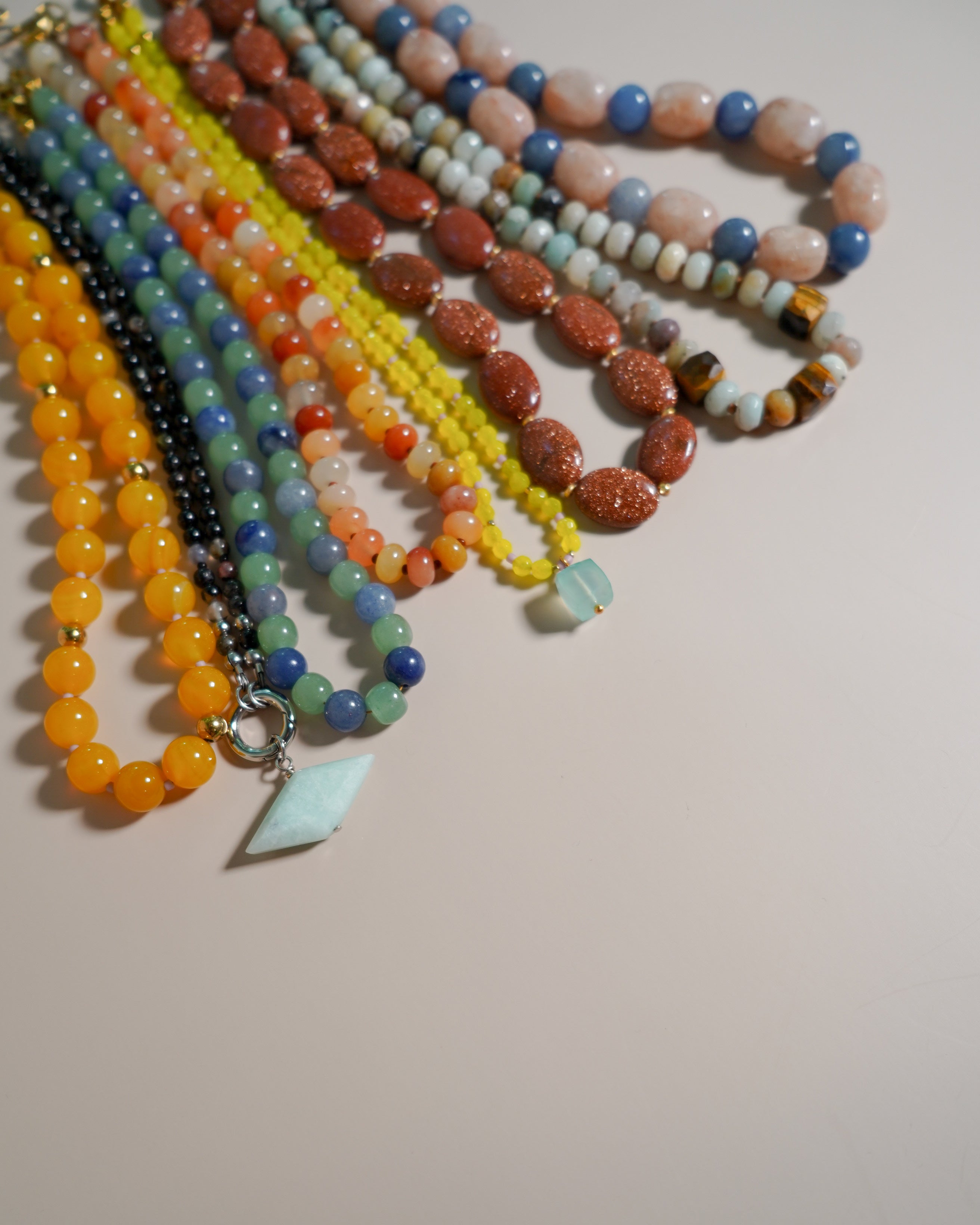 A collection of colorful beaded necklaces and bracelets arranged in a fan shape on a plain beige surface. The beads include various colors such as orange, yellow, blue, green, red, and brown, with one necklace featuring a light blue pendant.