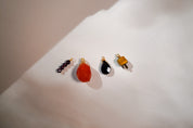 Four small jewelry charms or beads arranged on a white surface under warm directional light: a dark beaded pair, a faceted orange-red stone charm, a glossy black teardrop charm, and a small yellow-orange charm with gold-tone hardware.