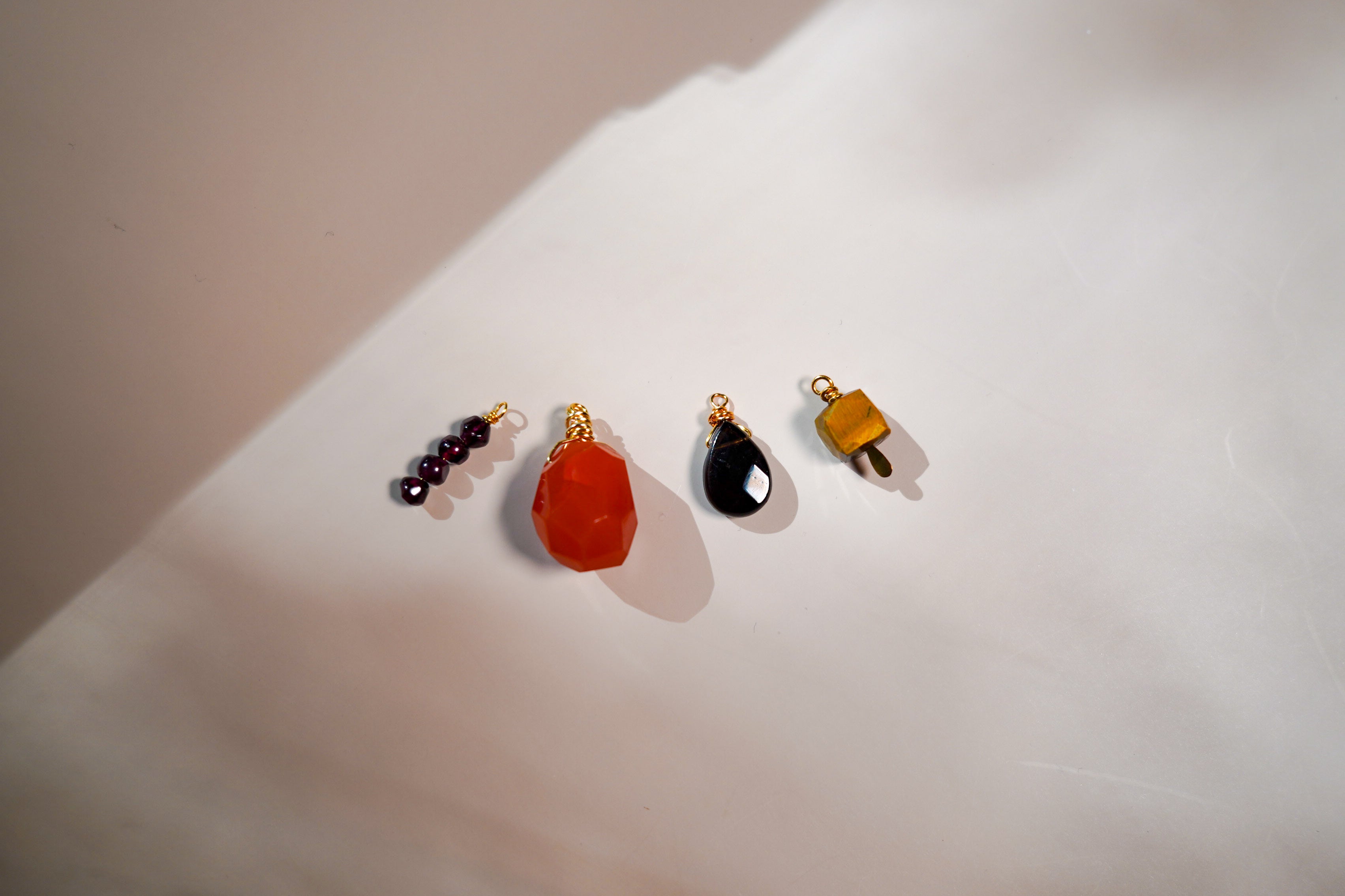 Four small jewelry charms or beads arranged on a white surface under warm directional light: a dark beaded pair, a faceted orange-red stone charm, a glossy black teardrop charm, and a small yellow-orange charm with gold-tone hardware.