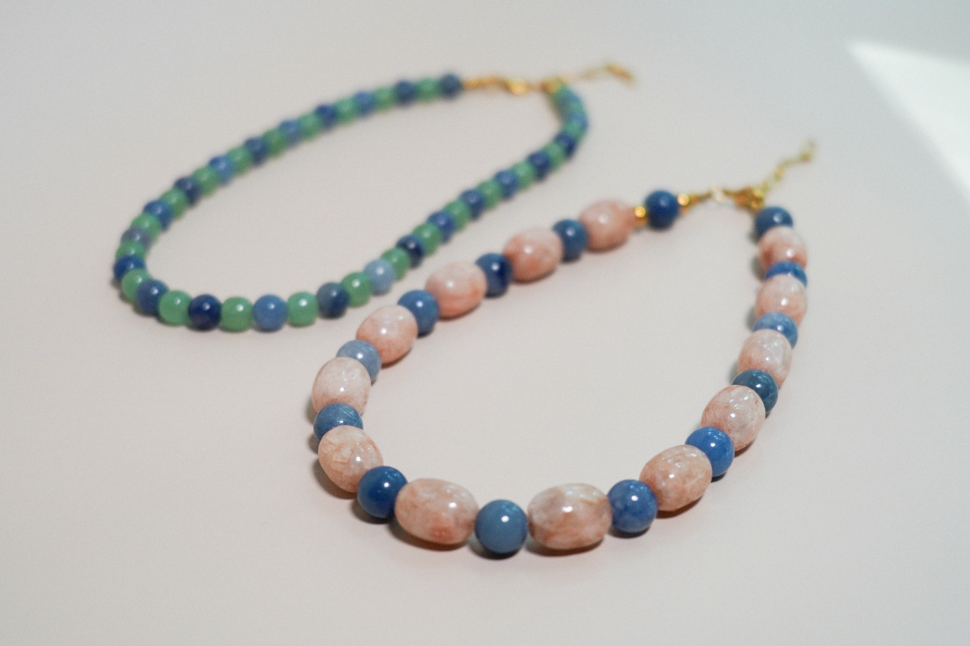 Two beaded necklaces displayed on a light beige surface. One necklace features round pinkish-orange and blue beads, while the other has green and blue beads, both with gold-tone clasp closures.