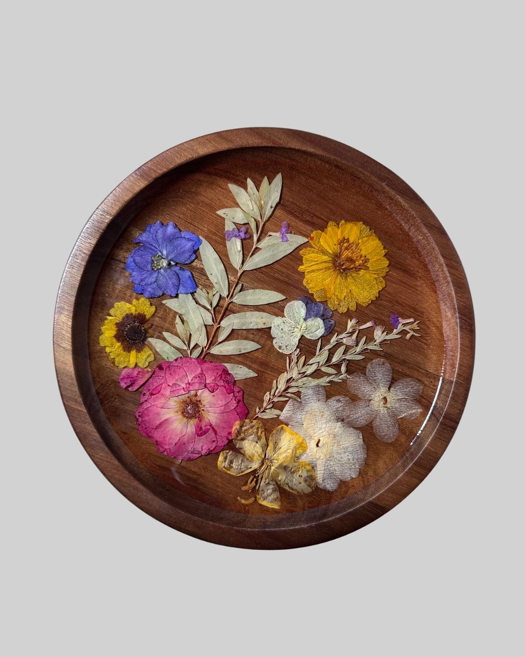 A round wooden tray with a raised rim, decorated with pressed and dried flowers and leaves in pink, yellow, blue, and white, set against a solid gray background.