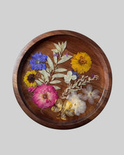 A round wooden tray with a raised rim, decorated with pressed and dried flowers and leaves in pink, yellow, blue, and white, set against a solid gray background.