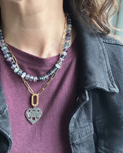A close-up of a person wearing layered necklaces. One necklace features light blue and purple beads with a silver heart-shaped pendant, and the other is a gold chain with a gold link pendant.
