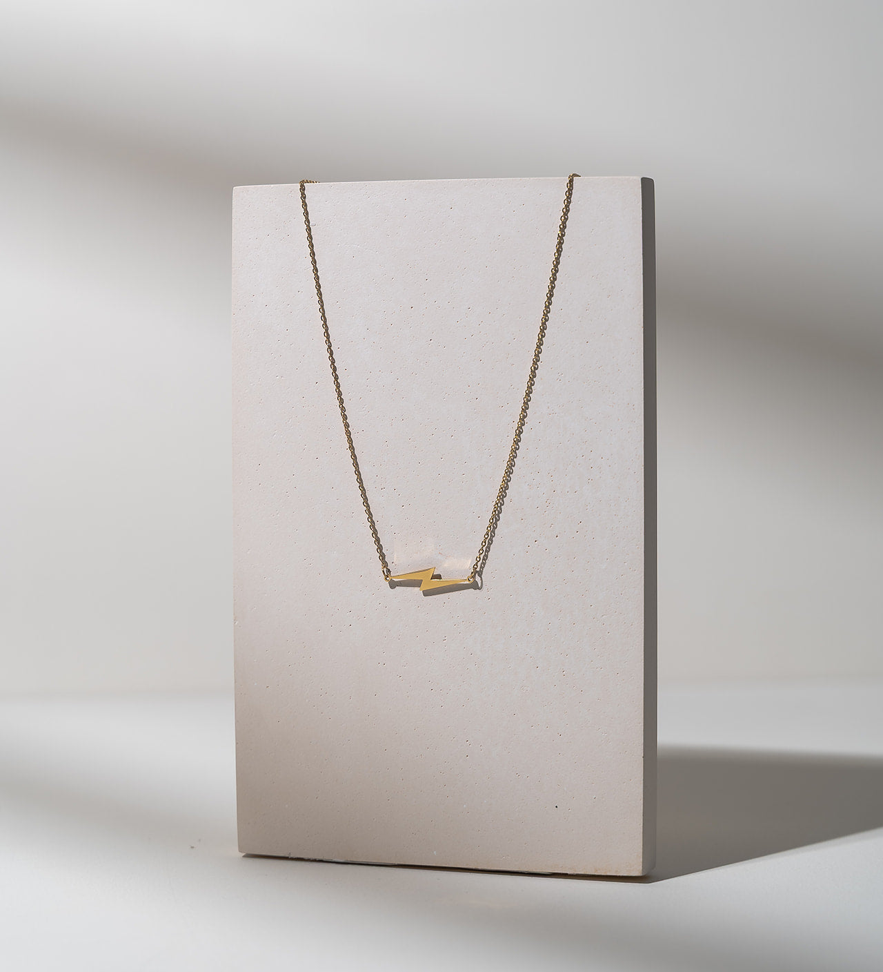 A delicate gold necklace with a minimalist bar pendant displayed on a textured, light beige vertical card against a neutral gray background.