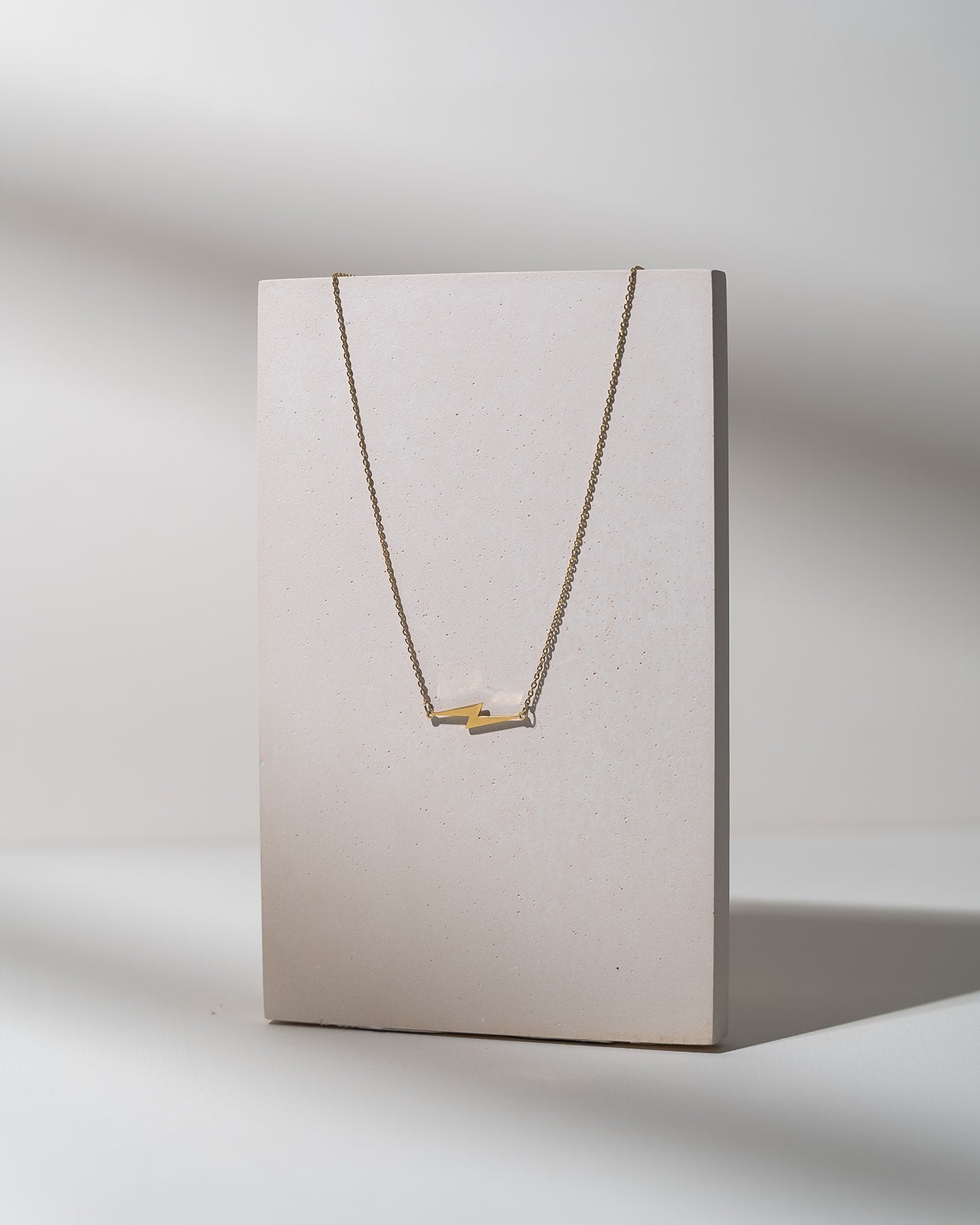 A delicate gold necklace with a minimalist bar pendant displayed on a textured, light beige vertical card against a neutral gray background.