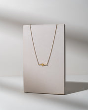 A delicate gold necklace with a minimalist bar pendant displayed on a textured, light beige vertical card against a neutral gray background.