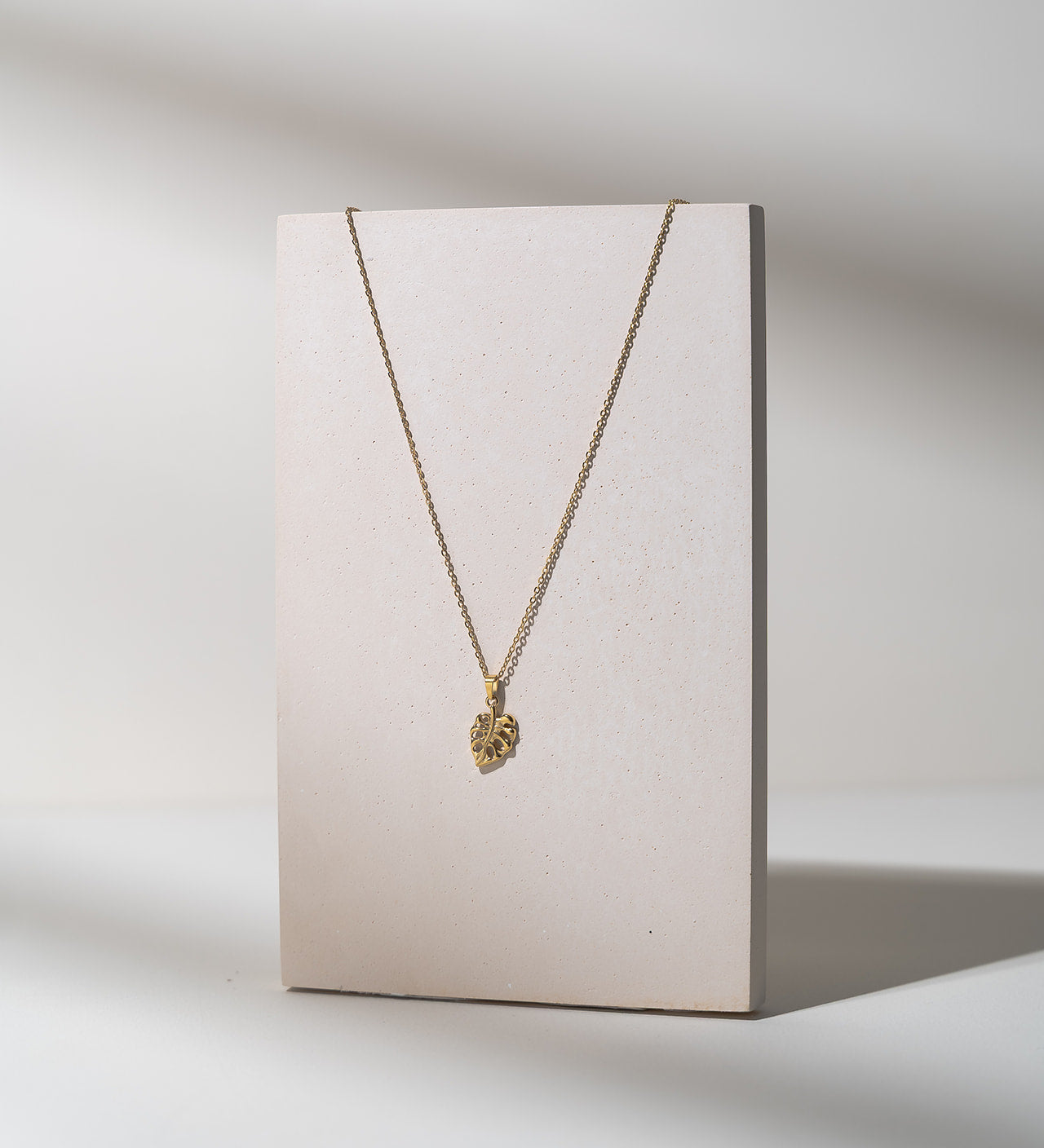 A delicate gold chain necklace with a small textured heart-shaped pendant, displayed on a minimalist beige rectangular stand against a clean white background with soft shadows.