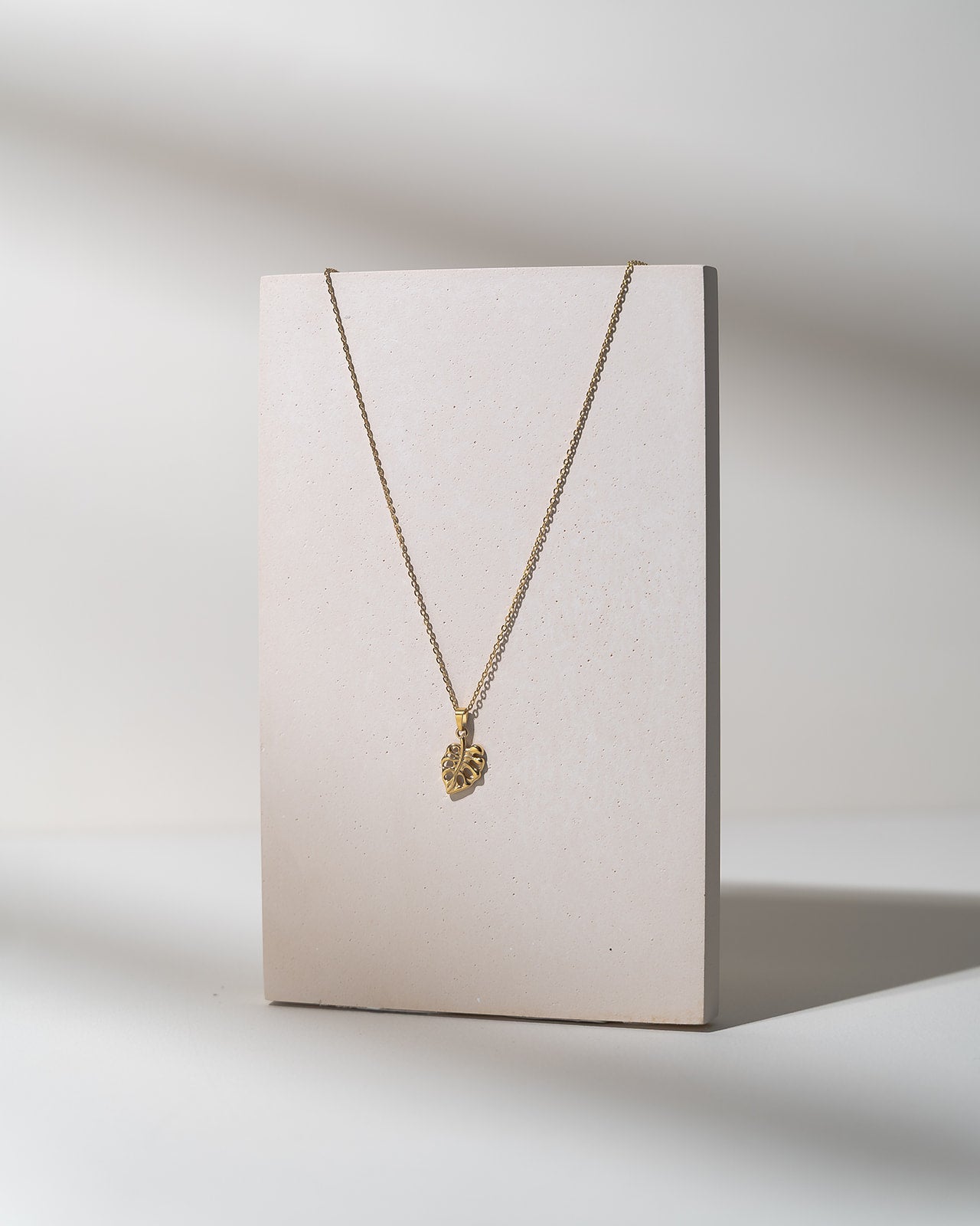 A delicate gold chain necklace with a small textured heart-shaped pendant, displayed on a minimalist beige rectangular stand against a clean white background with soft shadows.