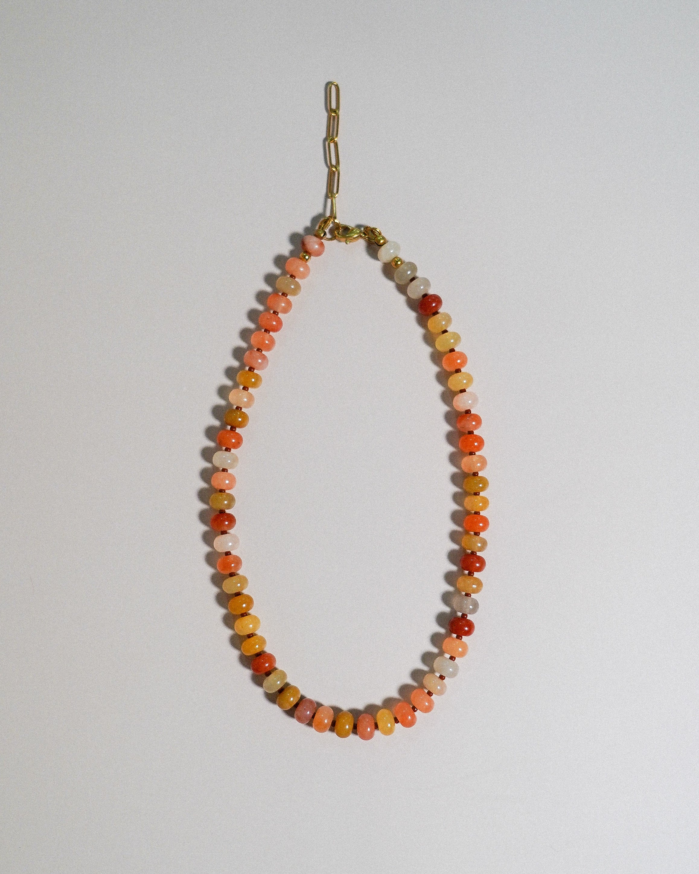 A beaded necklace made of small round stone beads in orange, peach, yellow, and cream tones, hanging from a gold-toned chain against a plain light background.