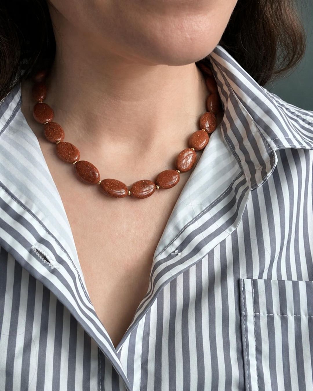 A close-up of a person wearing a beaded necklace made of polished reddish-brown oval beads, paired with a blue and white vertically striped button-down shirt.