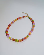 A beaded necklace made of small, round, translucent multicolor beads in shades of orange, yellow, green, pink, and red, with a gold clasp, displayed on a light gray background.
