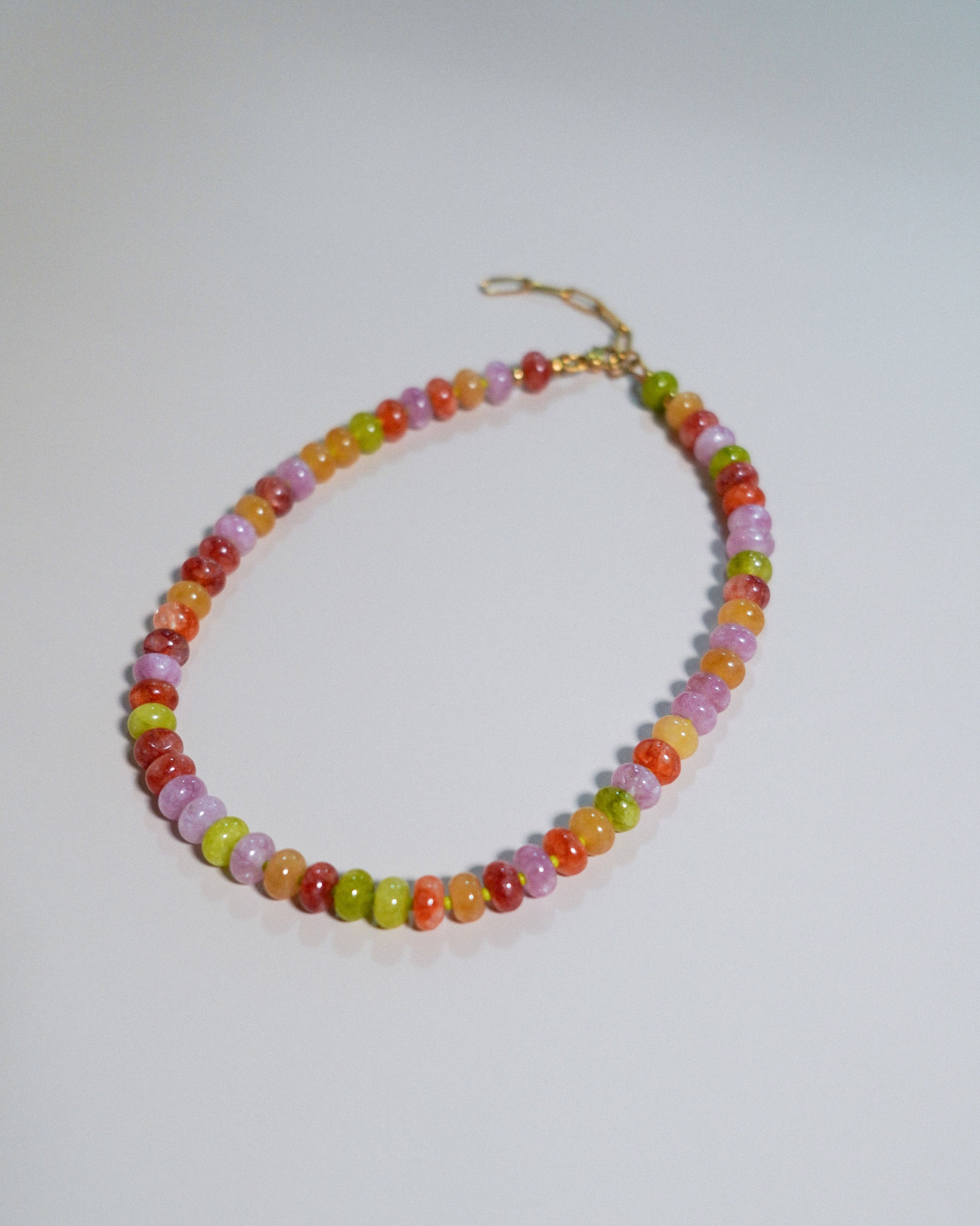 A beaded necklace made of small, round, translucent multicolor beads in shades of orange, yellow, green, pink, and red, with a gold clasp, displayed on a light gray background.