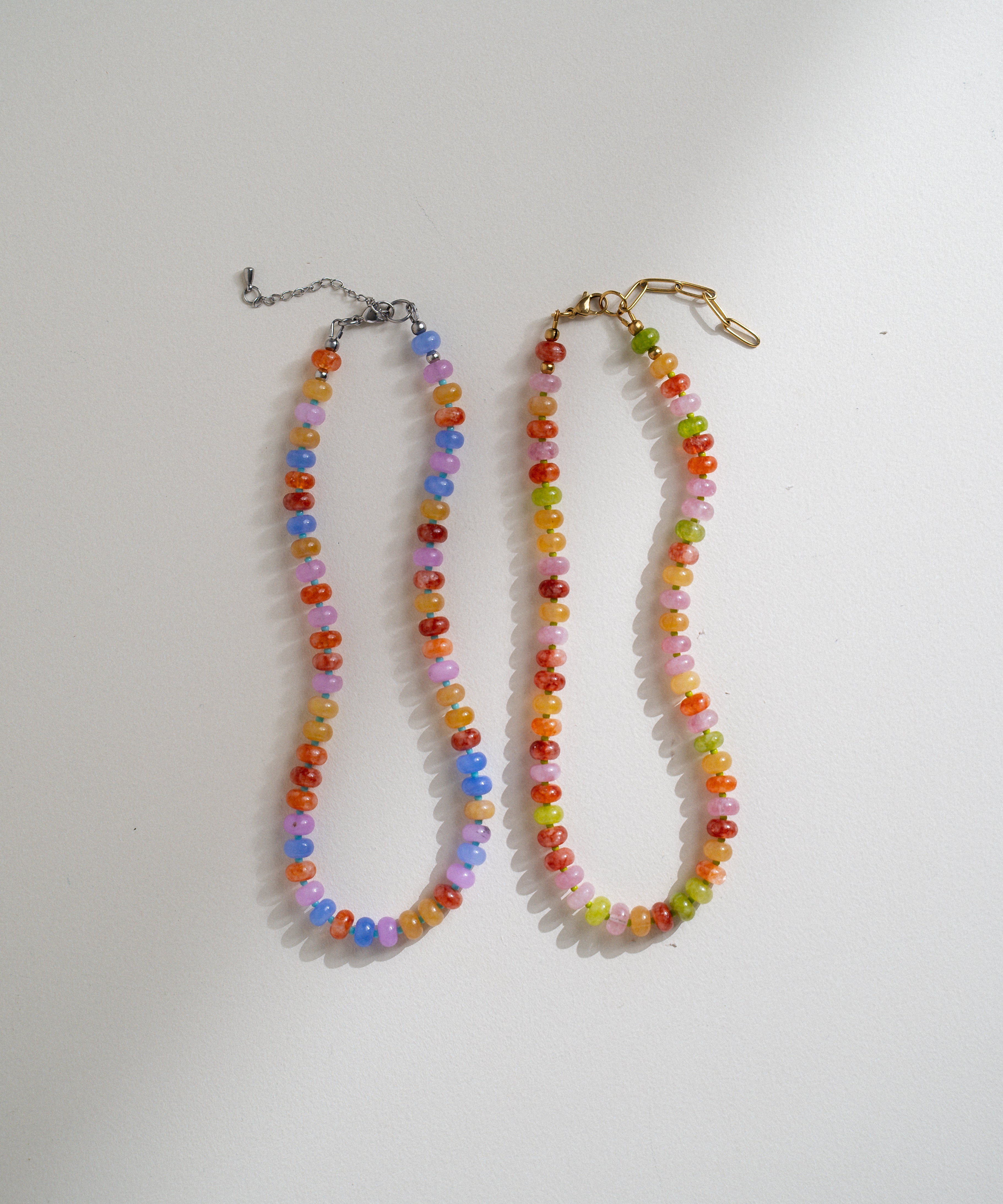 Two beaded necklaces laid flat on a white surface. One necklace has multicolored faceted beads with a silver-tone clasp and extender chain, and the other has multicolored beads with a gold-tone clasp and extender.