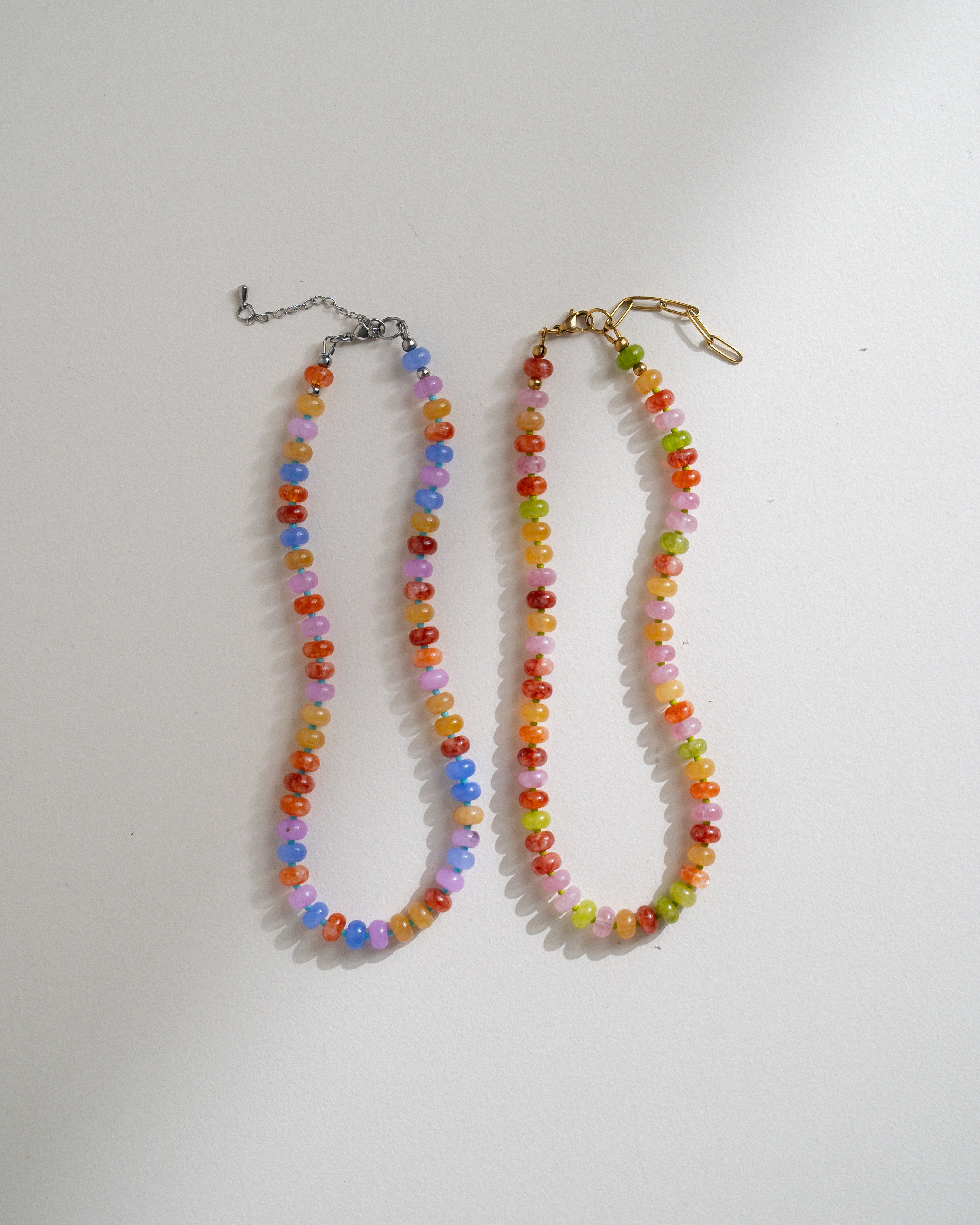Two beaded necklaces laid flat on a white surface. One necklace has multicolored faceted beads with a silver-tone clasp and extender chain, and the other has multicolored beads with a gold-tone clasp and extender.