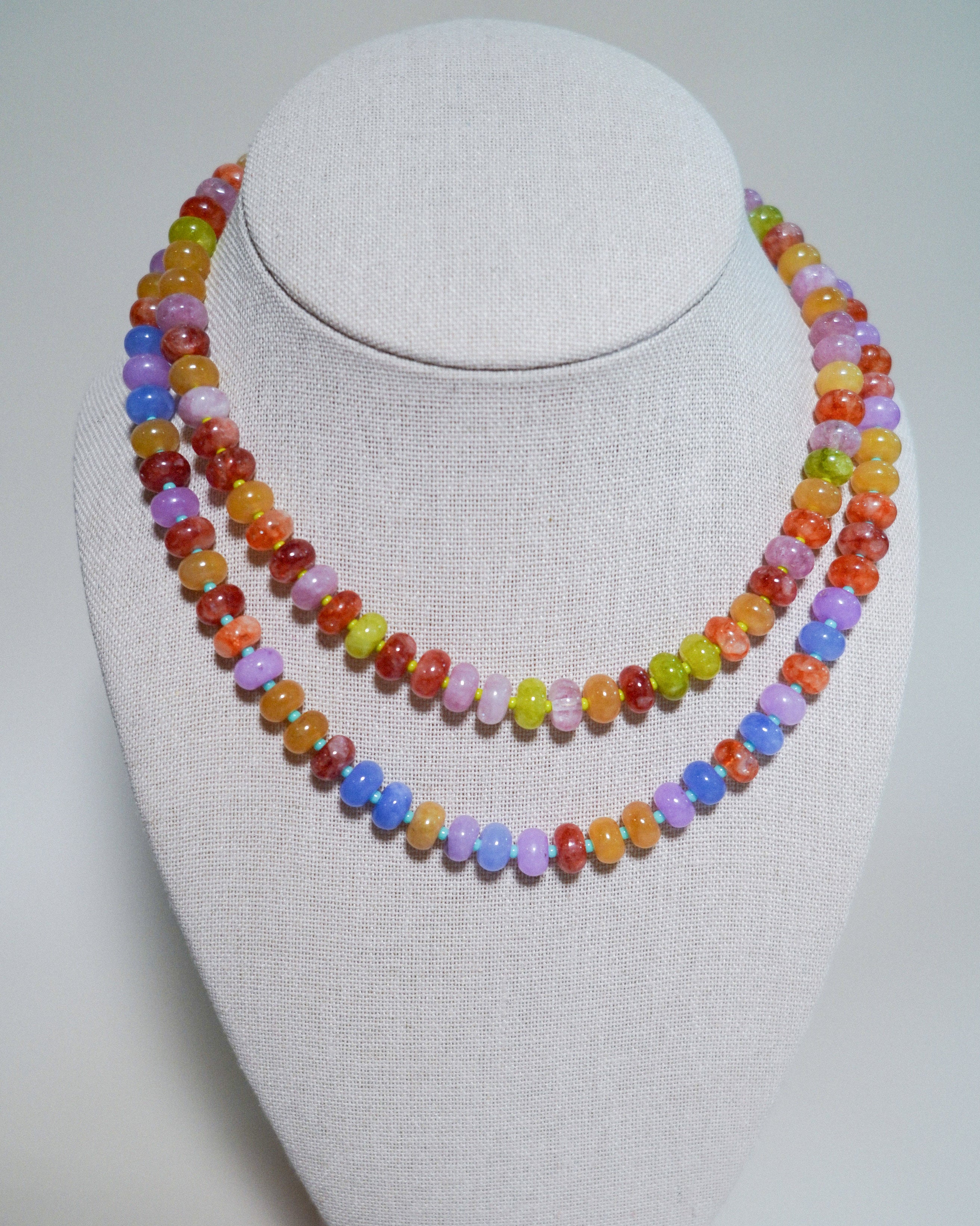 A double-strand beaded necklace displayed on a light gray fabric-covered jewelry bust. The necklace features faceted beads in multiple pastel and bright colors including orange, yellow, green, blue, purple, and red.
