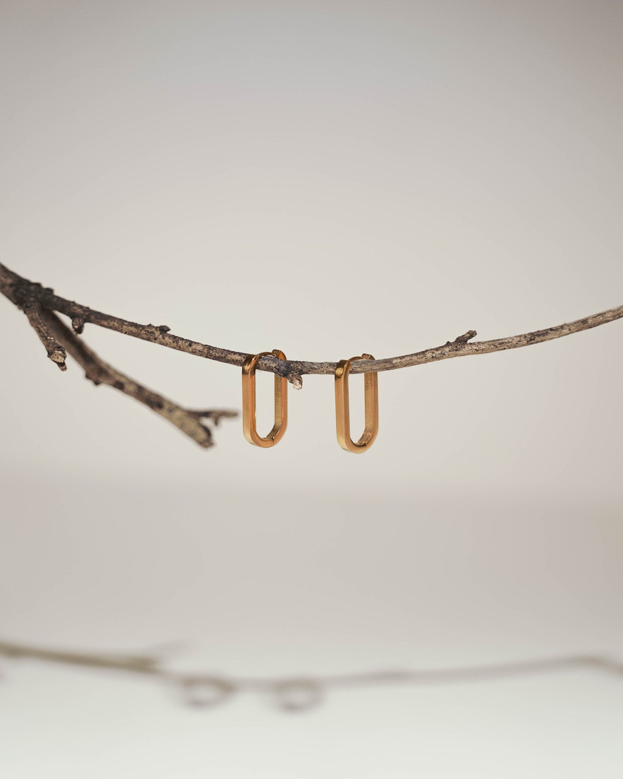 A pair of small gold U-shaped hoop earrings hanging from a thin natural twig against a soft light gray background.
