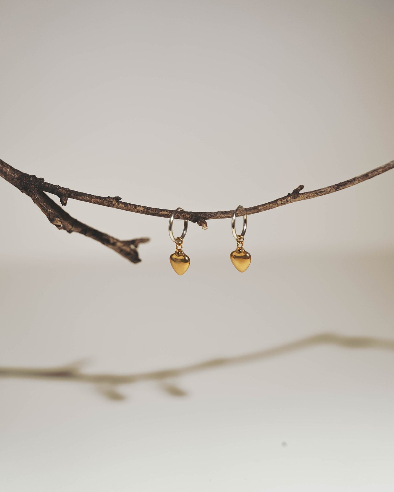 A pair of small gold hoop earrings with dangling heart-shaped charms, hanging from a thin brown branch against a soft light gray background.