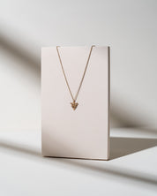 A delicate gold chain necklace with a small triangular pendant, displayed on a minimalist beige rectangular stand against a clean white background with soft shadows.