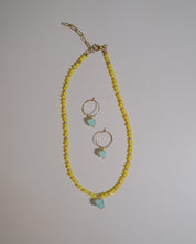 A gold-tone jewelry set featuring a beaded necklace and matching hoop earrings. The necklace is composed of small yellow faceted beads with a light blue stone accent at the bottom, while the matching hoop earrings have gold hoops and similar light blue stone drops.