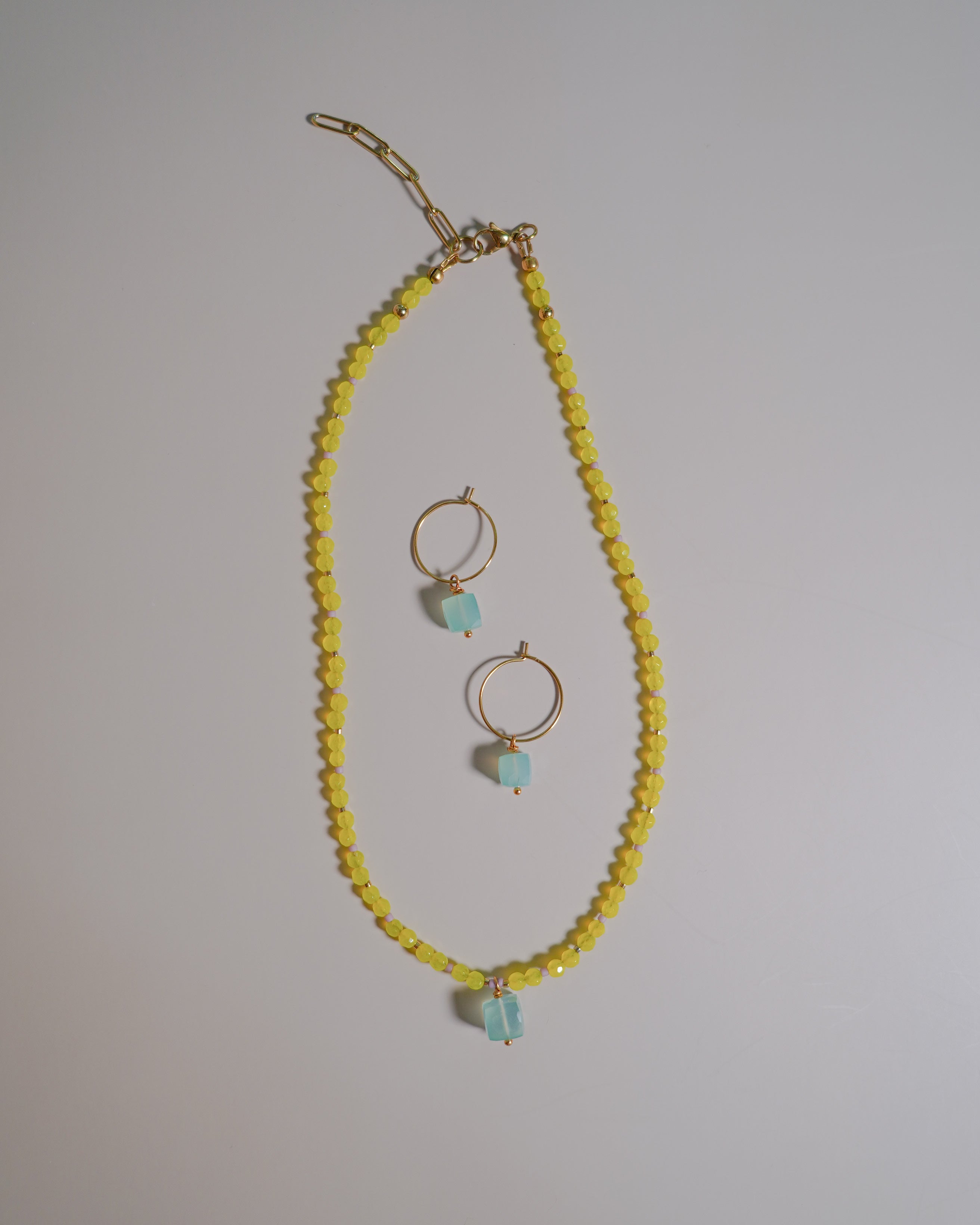 A gold-tone jewelry set featuring a beaded necklace and matching hoop earrings. The necklace is composed of small yellow faceted beads with a light blue stone accent at the bottom, while the matching hoop earrings have gold hoops and similar light blue stone drops.