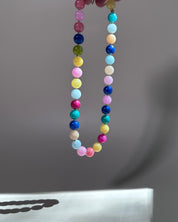 A beaded necklace made of small round pastel and bright gemstone-like beads in multiple colors, hanging against a soft gray background.