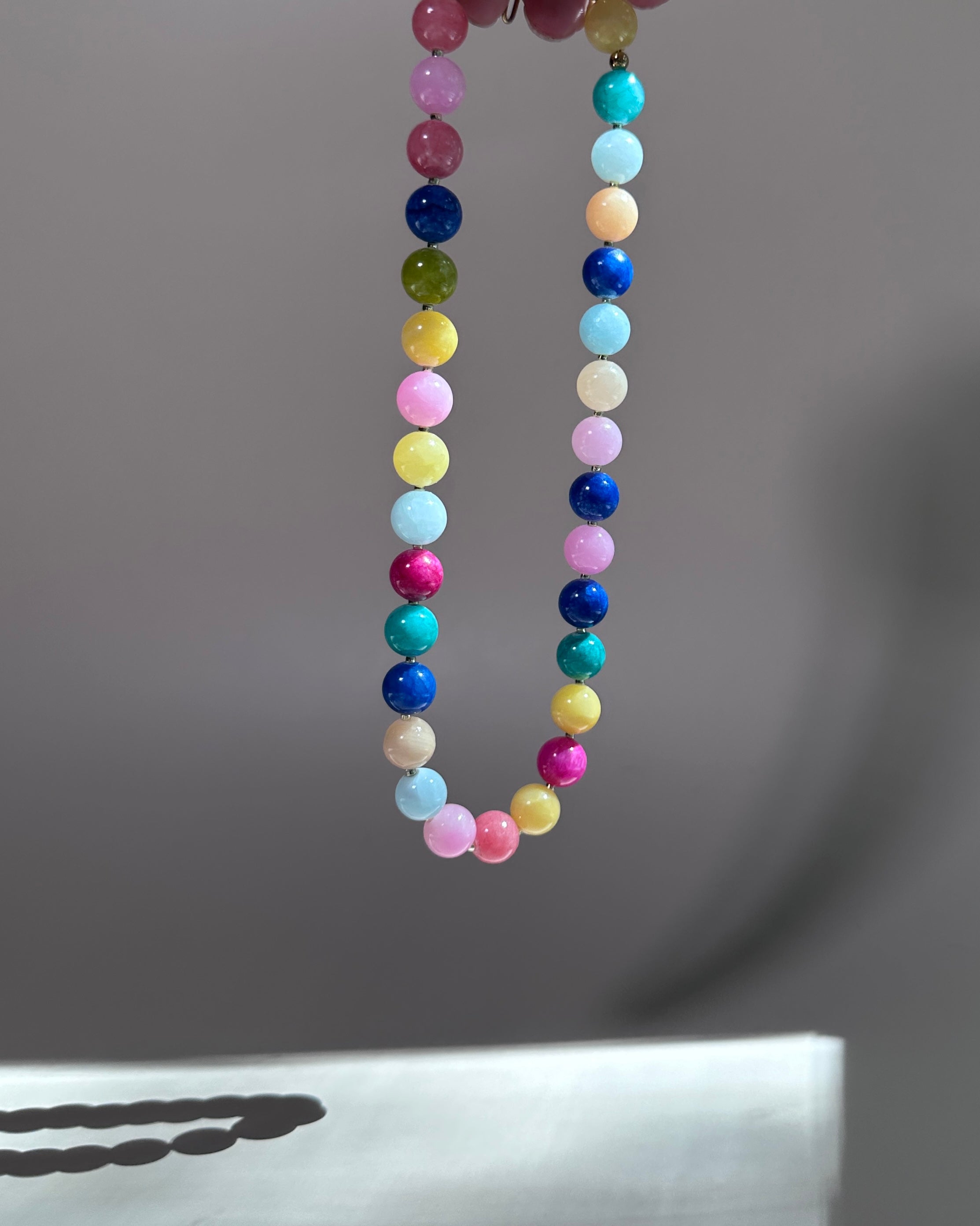 A beaded necklace made of small round pastel and bright gemstone-like beads in multiple colors, hanging against a soft gray background.