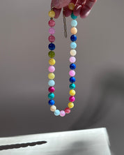 A beaded necklace with pastel and bright round beads in pink, yellow, blue, green, white, and orange, held by a hand against a neutral gray background.