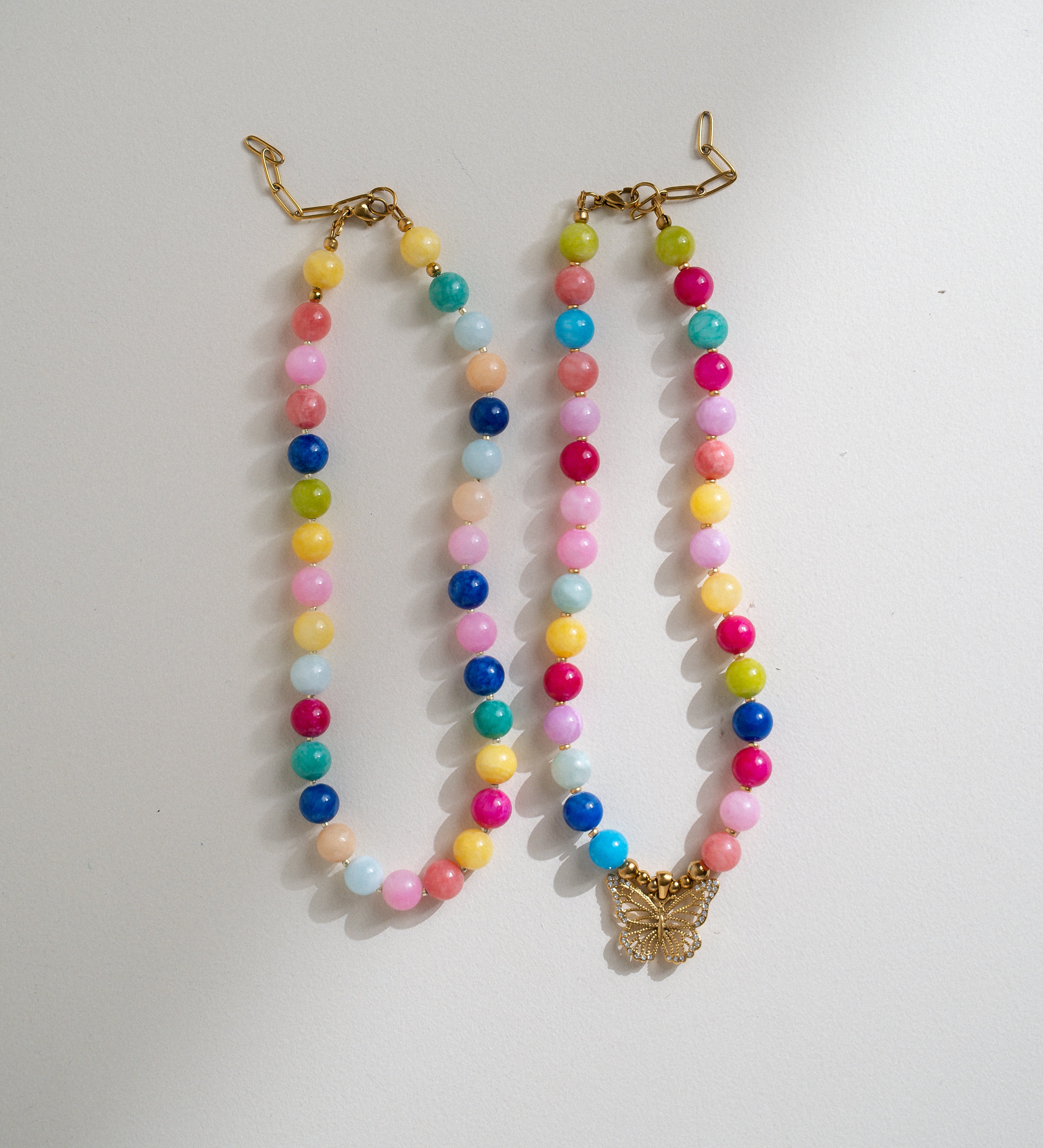 Two pastel beaded necklaces with gold-tone clasp closures and a butterfly pendant, displayed on a light gray background.