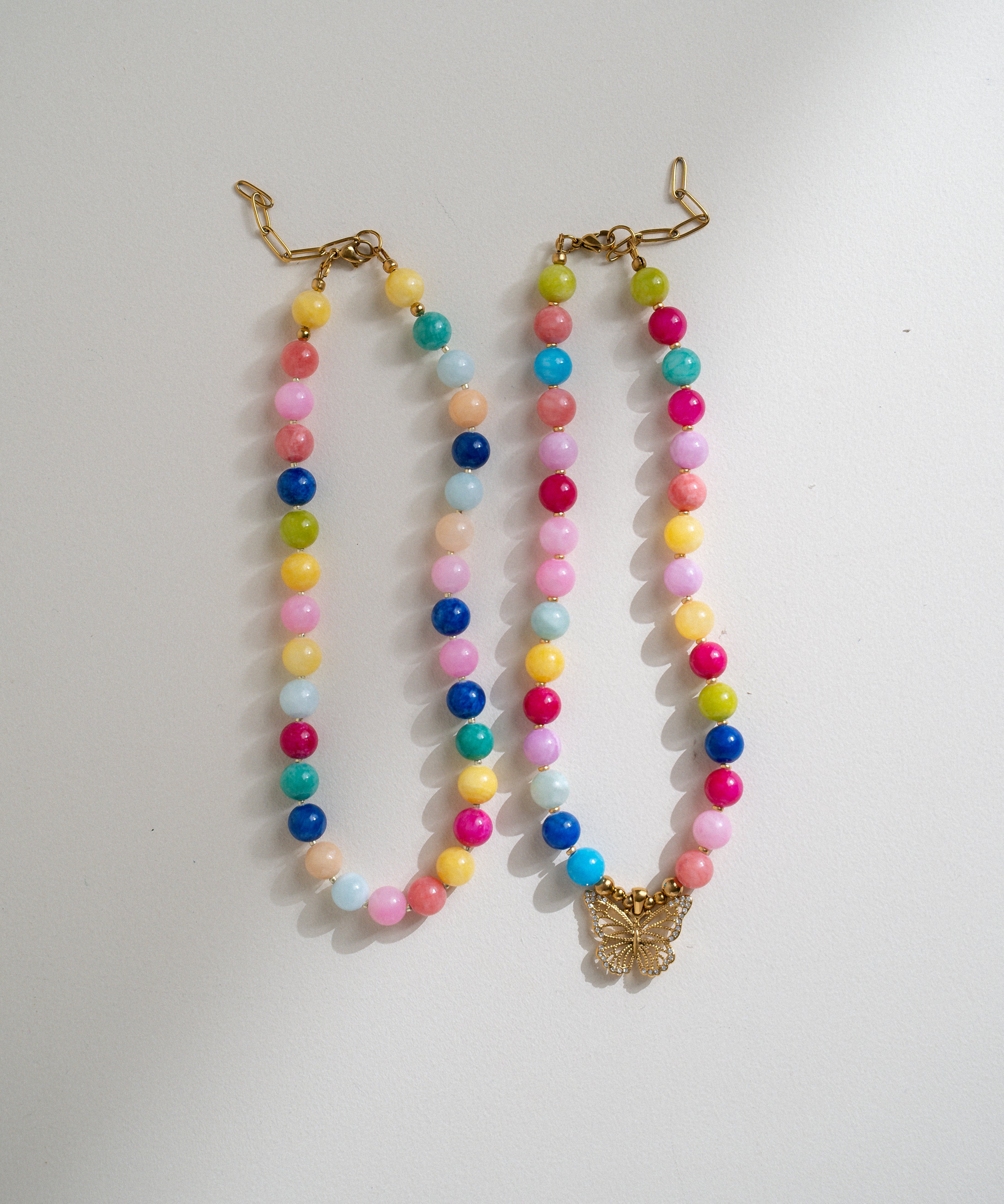 Two pastel beaded necklaces with gold-tone clasp closures and a butterfly pendant, displayed on a light gray background.