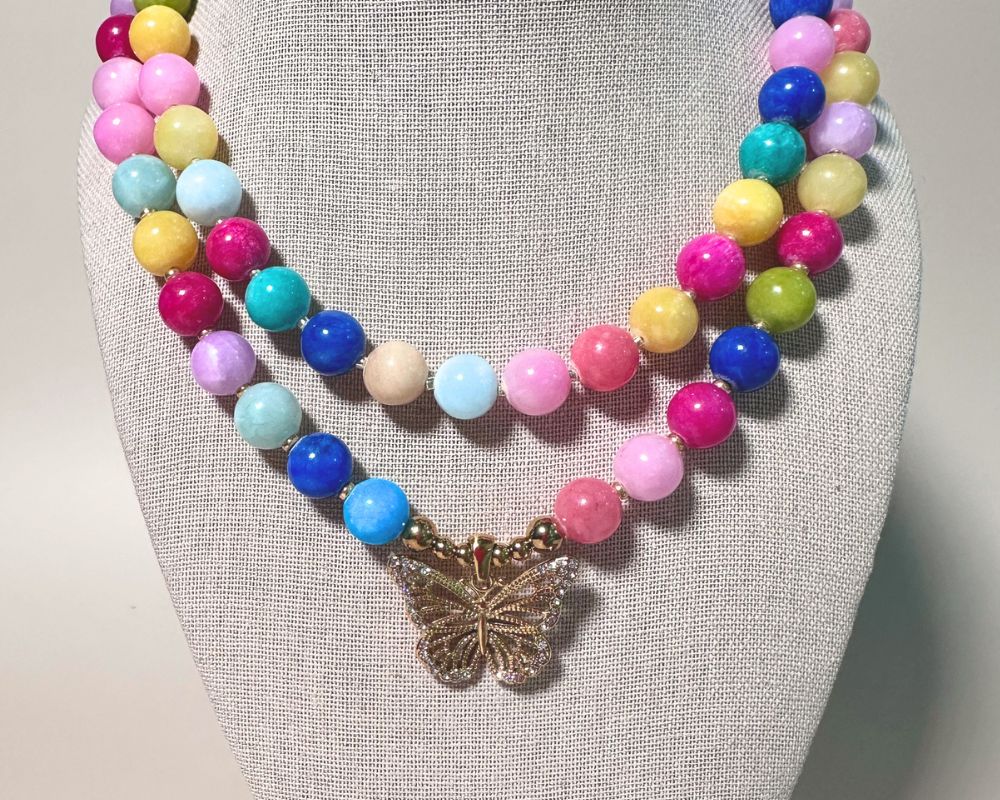 A multi-strand beaded necklace displayed on a neutral fabric mannequin bust. The necklace features pastel and bright multicolor round beads in pink, blue, yellow, green, and light blue, accented by a central gold butterfly charm.