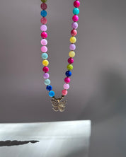 A colorful beaded necklace with a small butterfly pendant, hanging against a soft gray background.