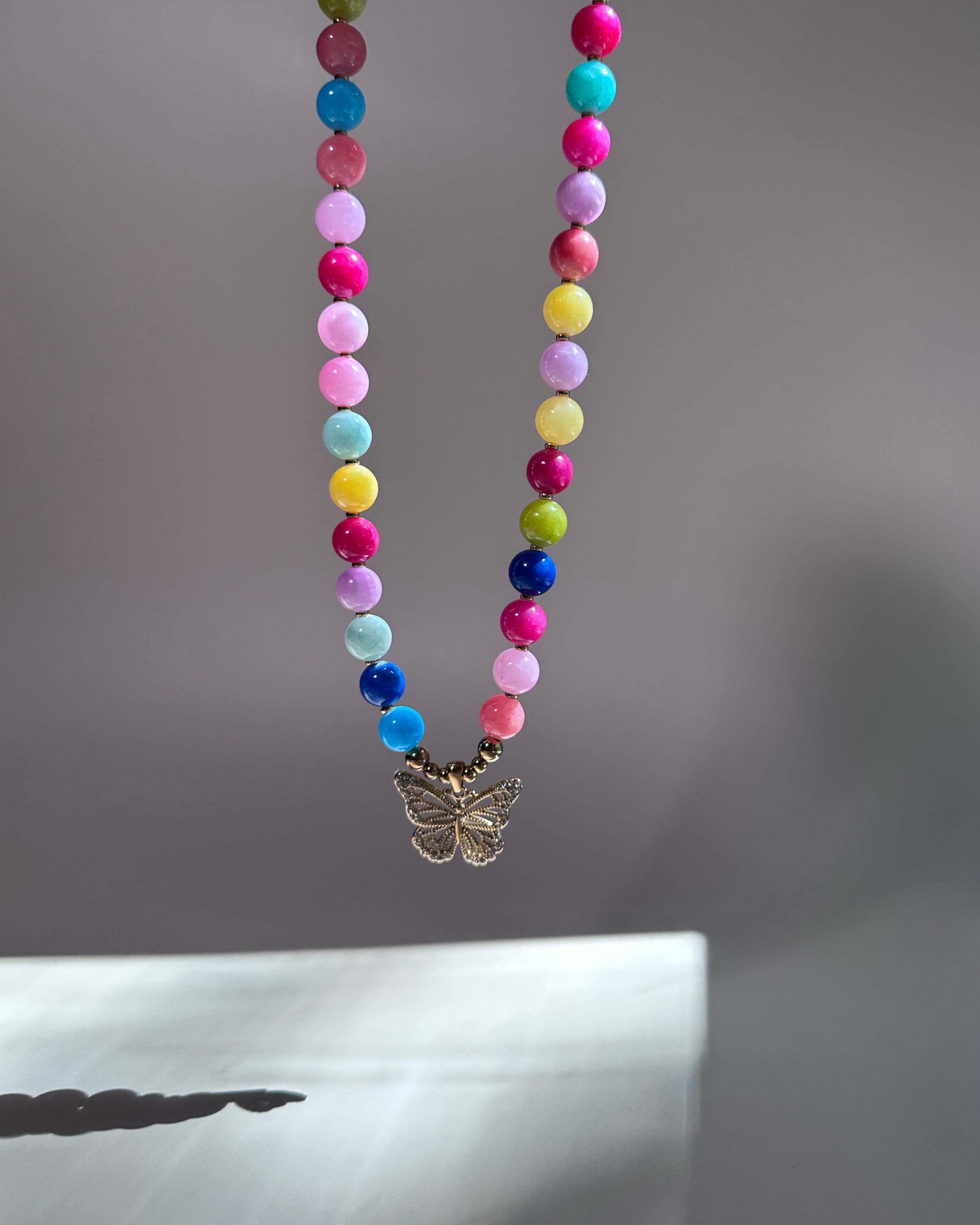 A colorful beaded necklace with a small butterfly pendant, hanging against a soft gray background.