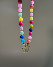 A colorful beaded necklace with round pastel and bright beads in pink, yellow, blue, green, purple, and white, hanging from a gold-tone butterfly pendant against a soft gray background.