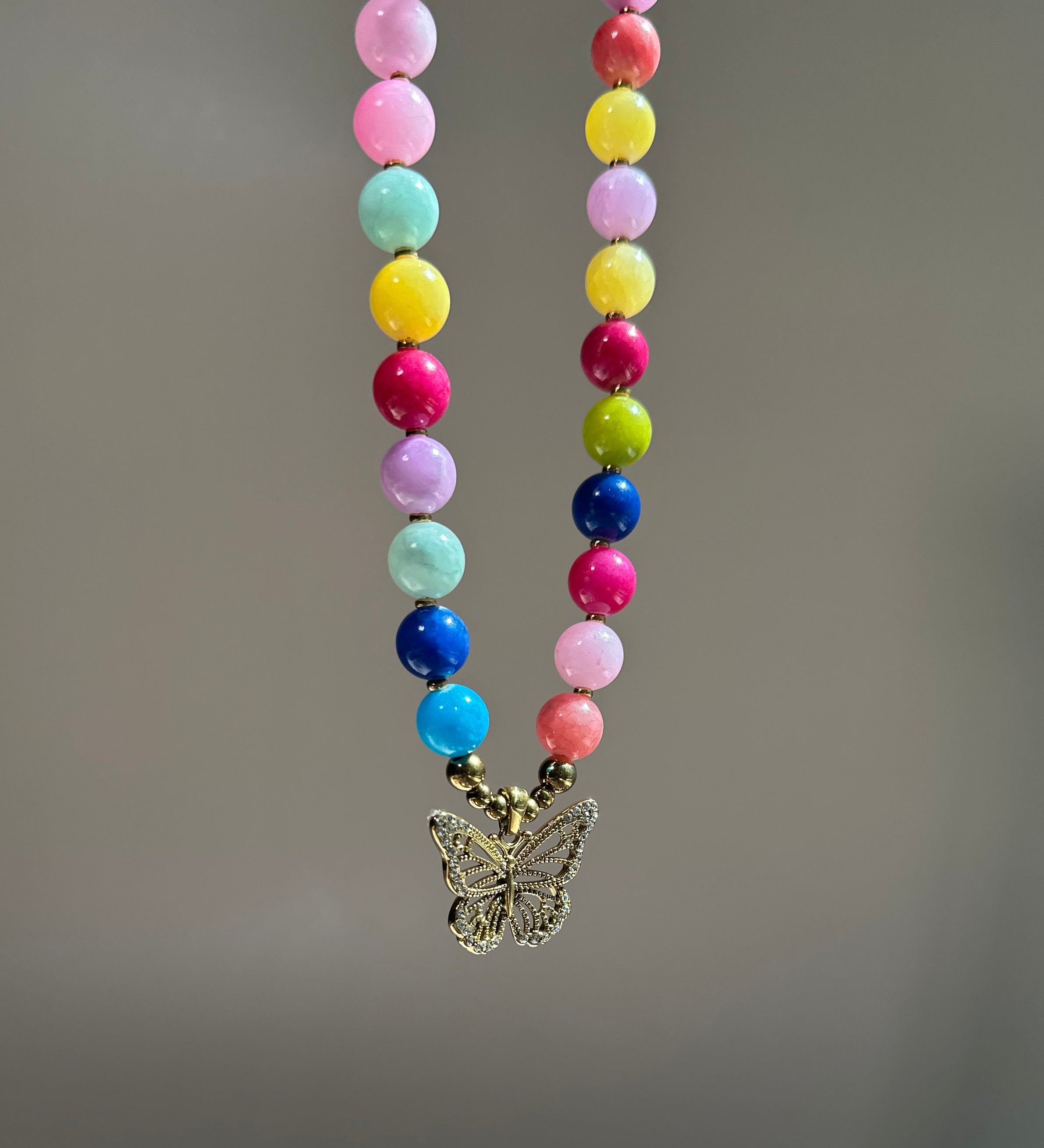 A colorful beaded necklace with round pastel and bright beads in pink, yellow, blue, green, purple, and white, hanging from a gold-tone butterfly pendant against a soft gray background.