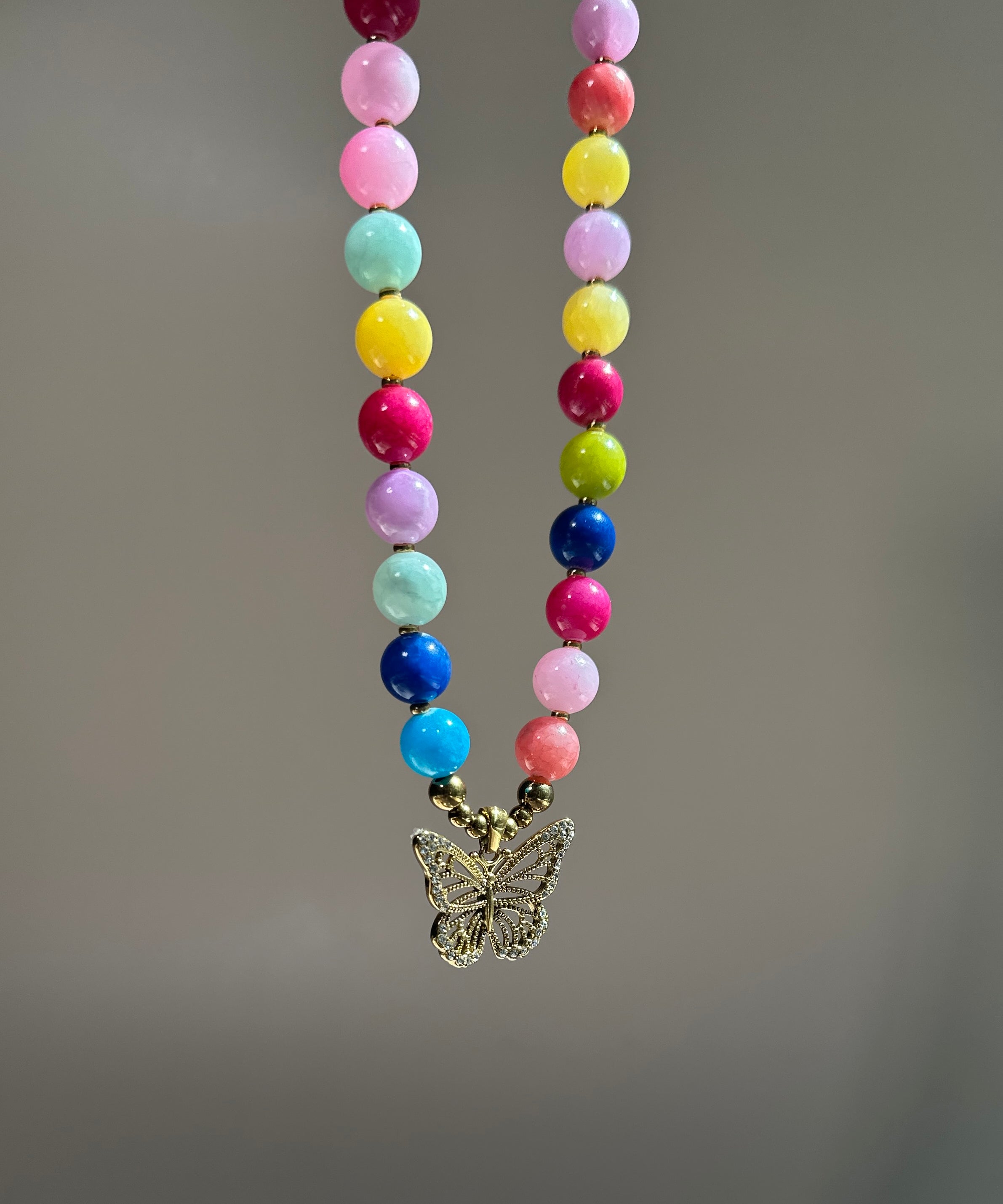 A colorful beaded necklace with round pastel and bright beads in pink, yellow, blue, green, purple, and white, hanging from a gold-tone butterfly pendant against a soft gray background.
