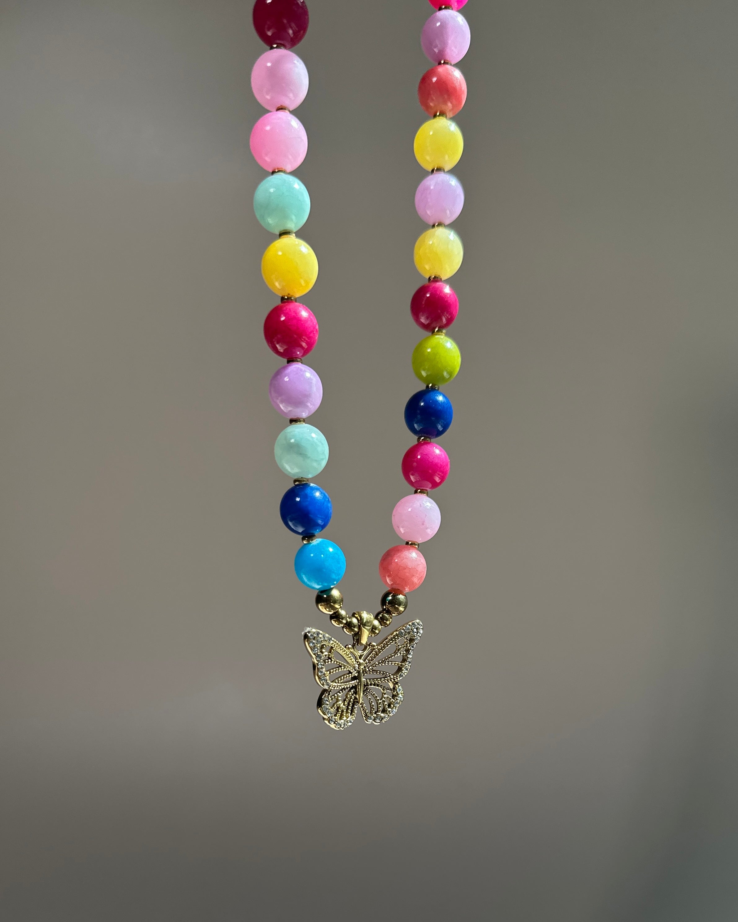 A colorful beaded necklace with round pastel and bright beads in pink, yellow, blue, green, purple, and white, hanging from a gold-tone butterfly pendant against a soft gray background.