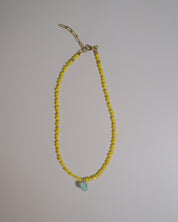 A delicate beaded necklace featuring small yellow faceted beads and a small light blue teardrop-shaped bead at the center, suspended from a gold-toned chain clasp against a plain light gray background.