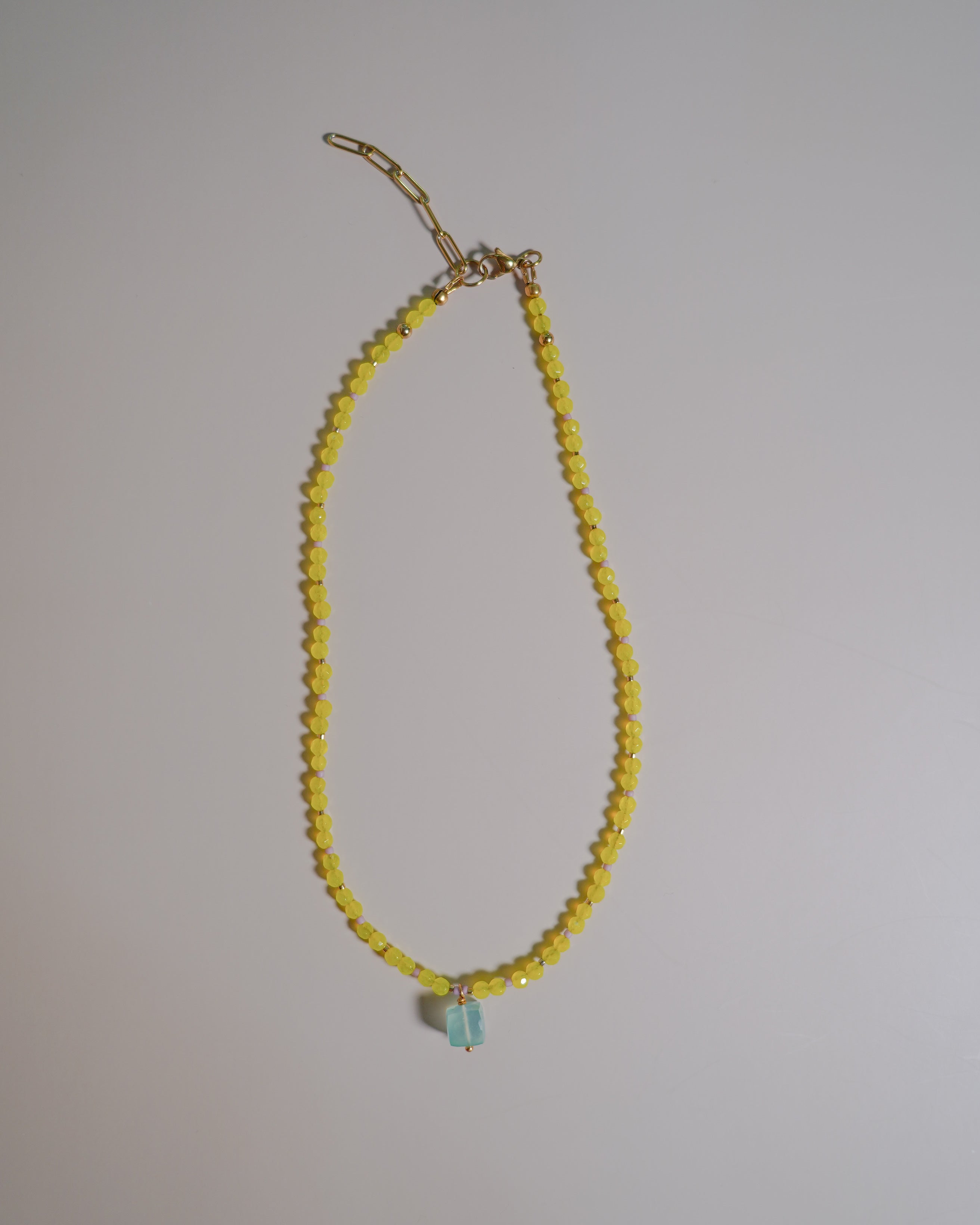 A delicate beaded necklace featuring small yellow faceted beads and a small light blue teardrop-shaped bead at the center, suspended from a gold-toned chain clasp against a plain light gray background.