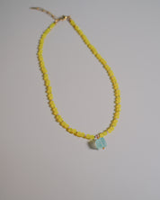 A beaded necklace featuring a strand of small yellow faceted beads and a central light blue faceted gemstone pendant, displayed on a neutral light gray background.