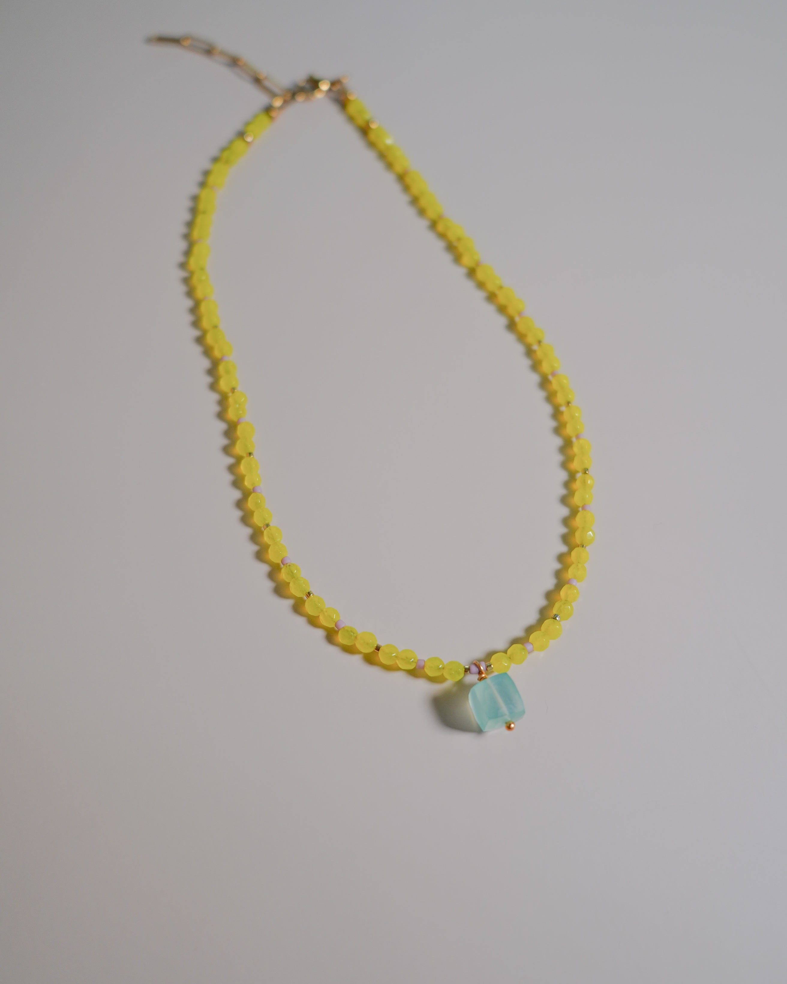 A beaded necklace featuring a strand of small yellow faceted beads and a central light blue faceted gemstone pendant, displayed on a neutral light gray background.