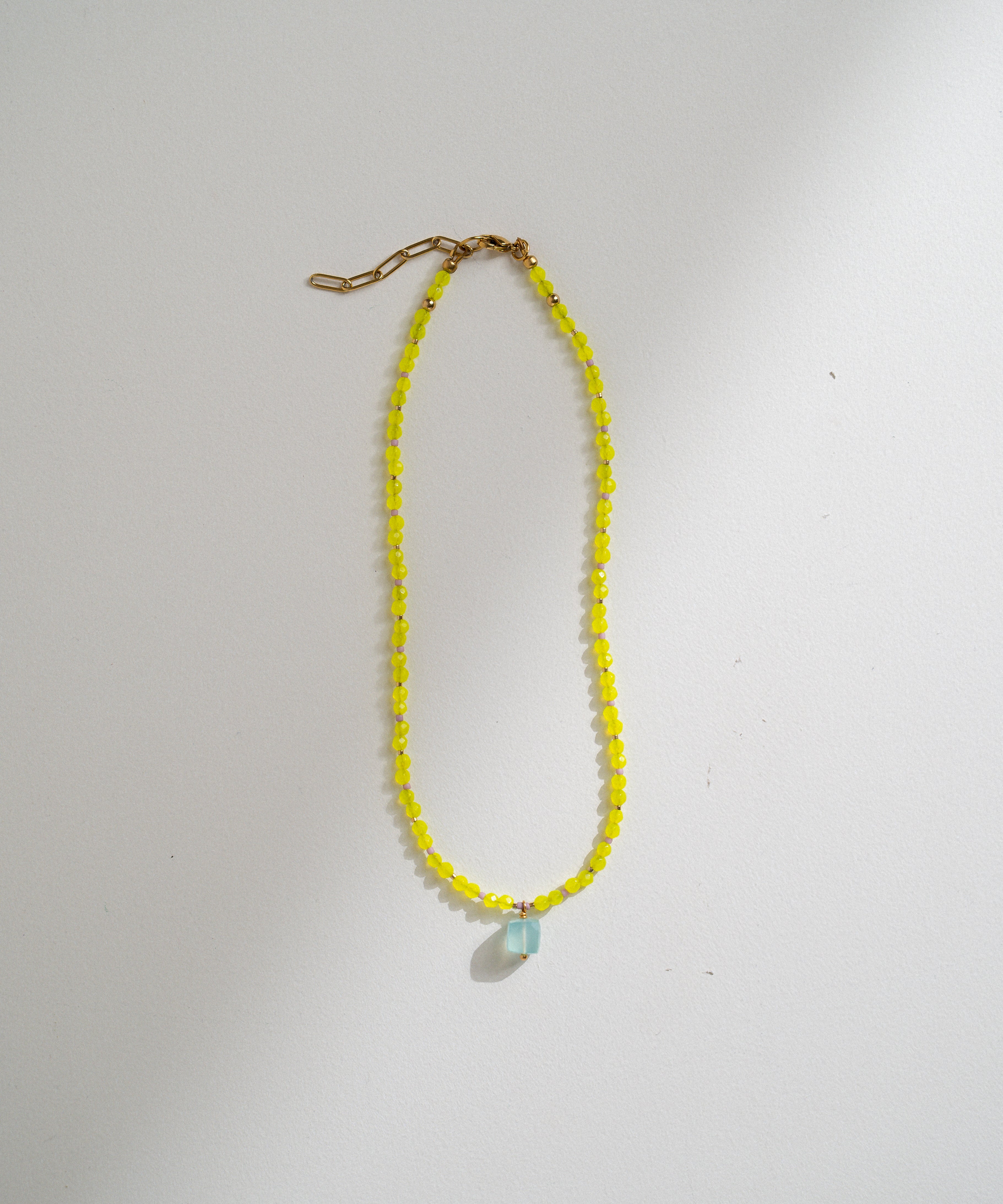 A delicate necklace made of small yellow faceted beads with a small light blue teardrop pendant, hanging against a plain off-white background.