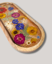 An oval wooden tray with a raised rim, decorated with pressed dried flowers in yellow, pink, blue, and white on a light tan background, shown on a solid gray background.