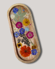 An oval wooden tray with a raised rim, decorated with pressed wildflowers and green leaves under a clear resin cover, set against a solid light gray background.