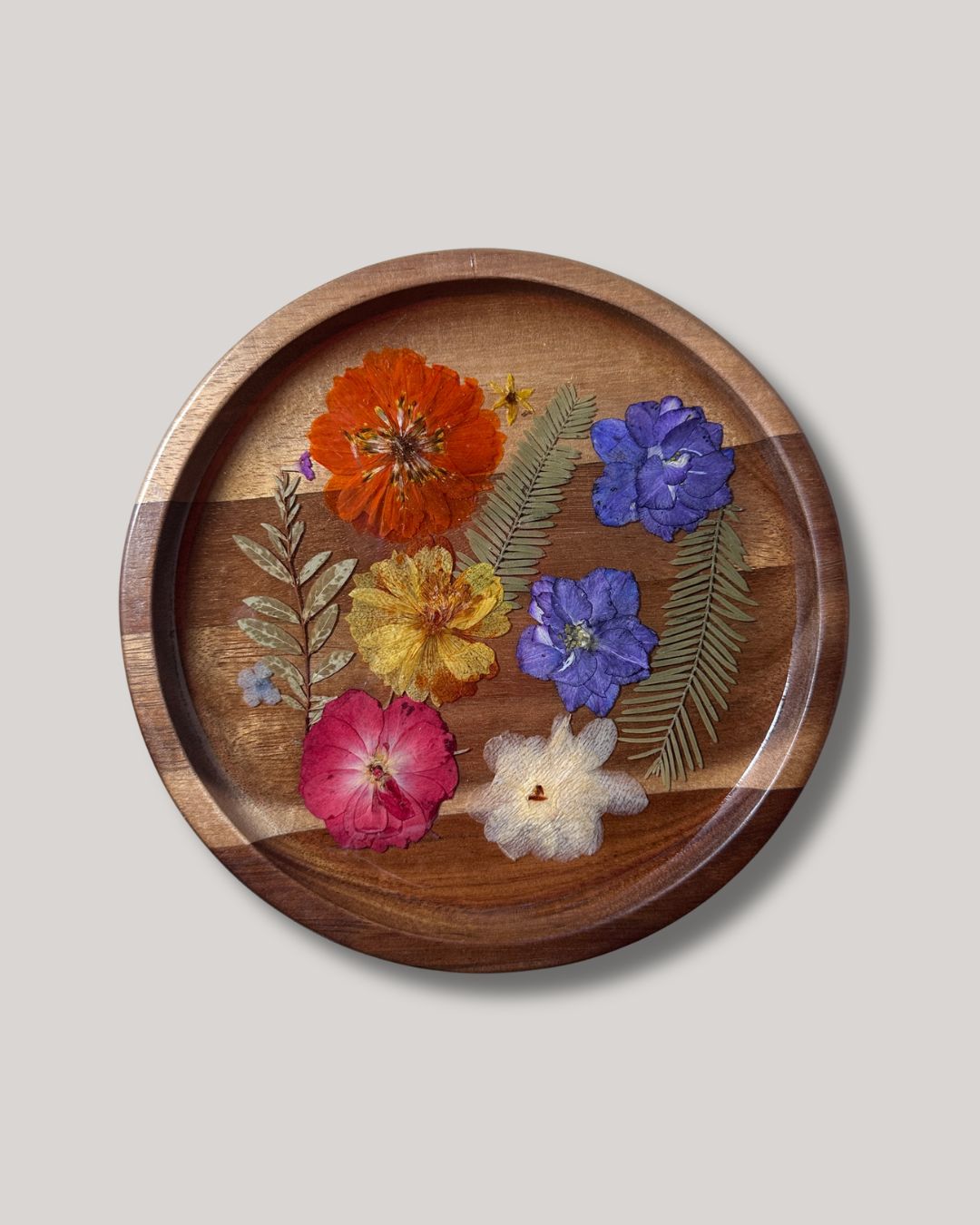 A round wooden tray featuring a colorful arrangement of pressed dried flowers and green leaves, set against a solid light gray background.