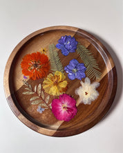 A round wooden tray with a raised rim, decorated with pressed flowers and green leaves in orange, purple, yellow, pink, and white, photographed from above on a light neutral background.