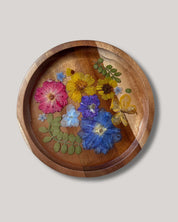 A round wooden tray with a carved floral design featuring pressed flowers in pink, yellow, blue, and purple with green leaves, set against a solid light gray background.